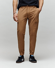 A person wearing Psycho Bunny MENS LLOYD CARGO JOGGER - B6P159B200 with side pockets and subtle embroidery, paired with a black t-shirt and black Adidas sneakers with white stripes, stands against a plain gray background.