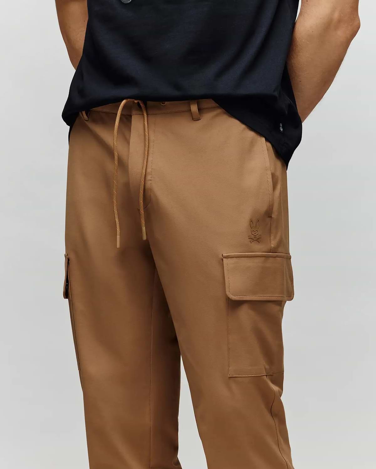 A person wears Psycho Bunny MENS LLOYD CARGO JOGGER - B6P159B200, tan cargo joggers with drawstring waist, side pockets, and subtle embroidery, paired with a black shirt and hands tucked behind their back.