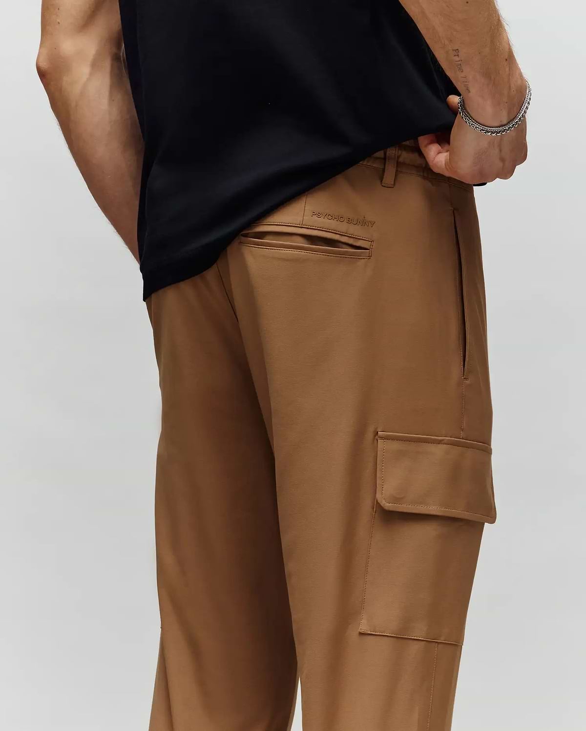 A person wears Psycho Bunny MENS LLOYD CARGO JOGGER - B6P159B200 in light brown, shown from the waist down with one hand in their pocket. The joggers have a back pocket, side cargo pocket, and subtle embroidery detail.