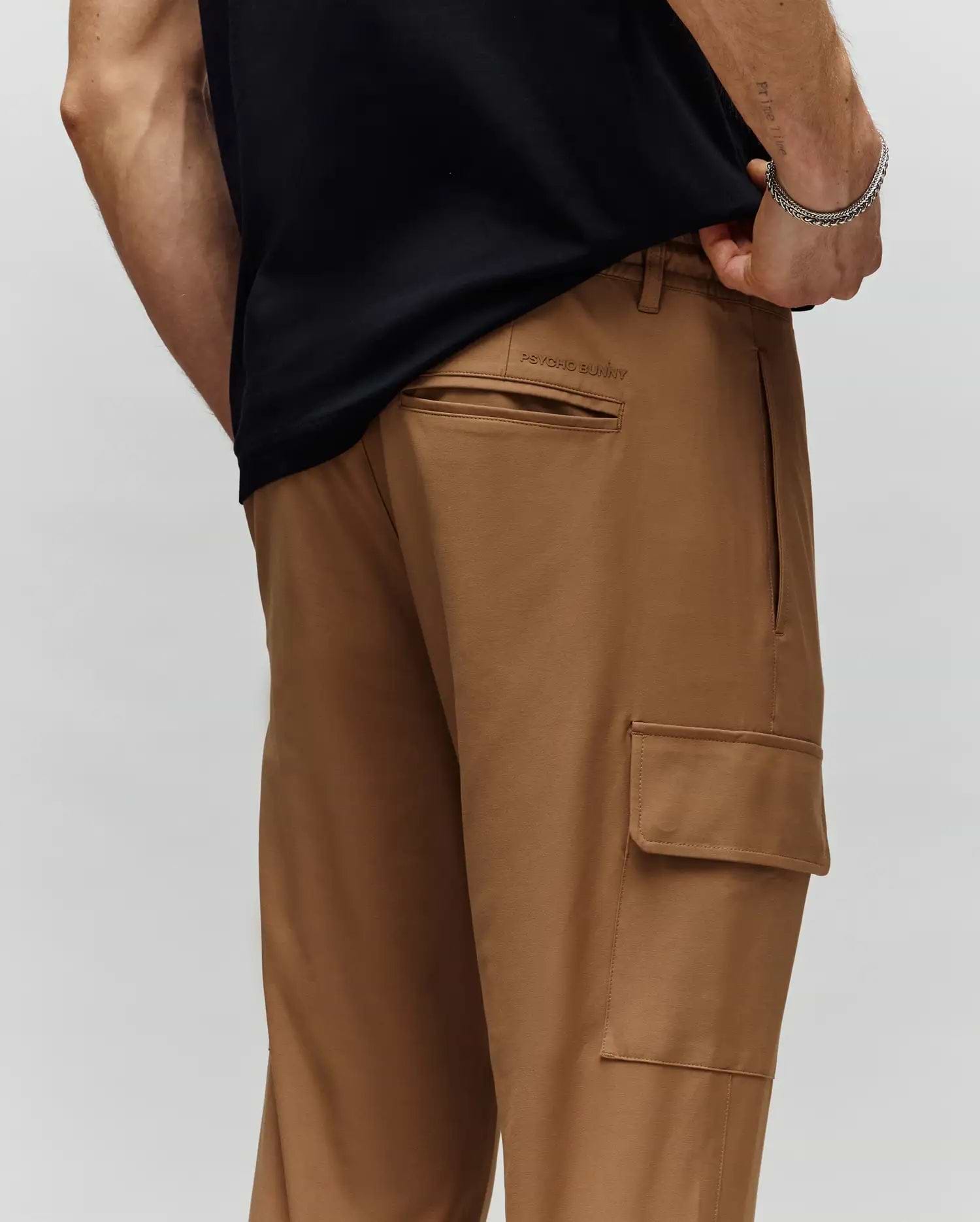 A person wears Psycho Bunny MENS LLOYD CARGO JOGGER - B6P159B200 in light brown, shown from the waist down with one hand in their pocket. The joggers have a back pocket, side cargo pocket, and subtle embroidery detail.
