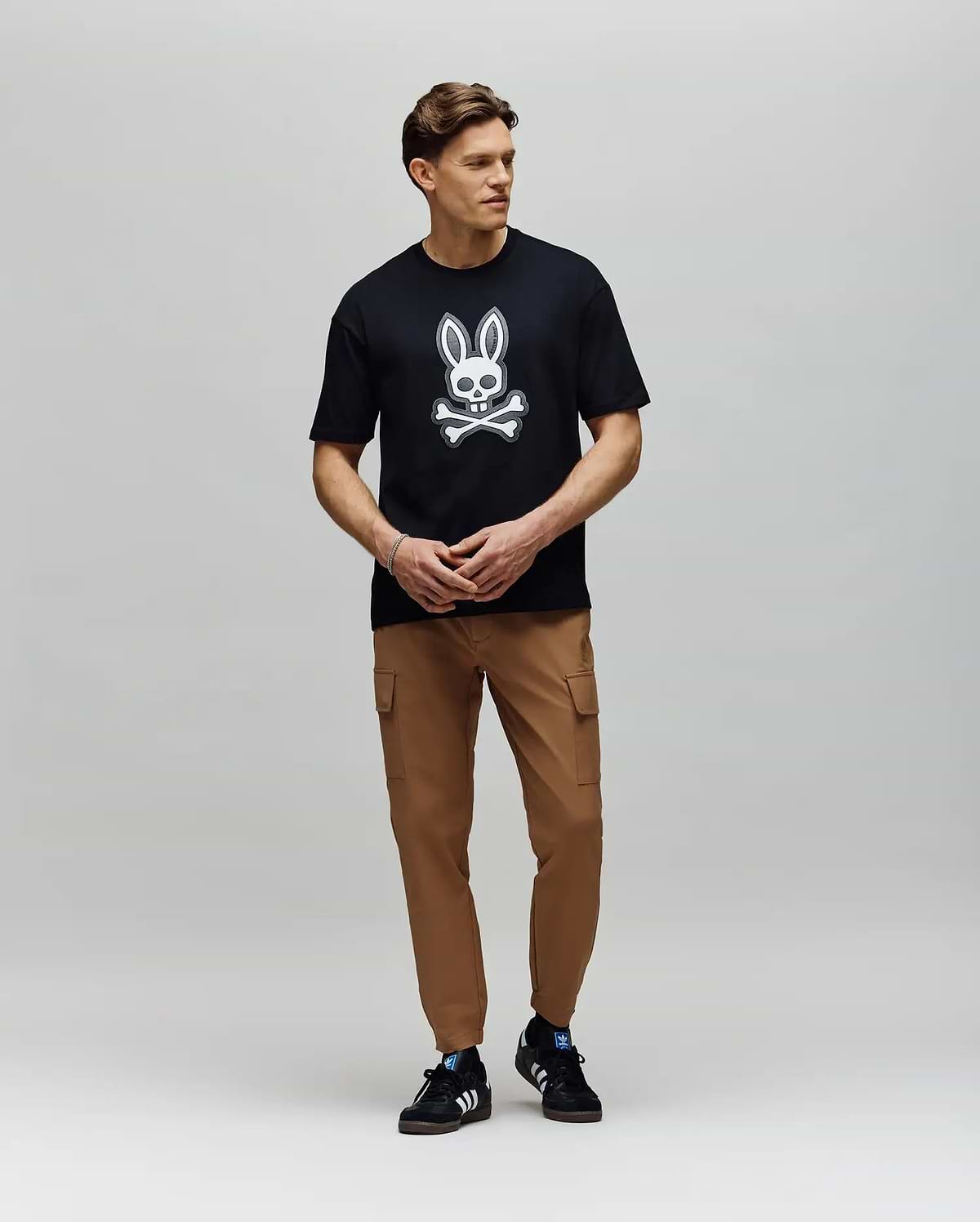 A man stands against a plain background wearing a black Psycho Bunny T-shirt with a white bunny and crossbones, MENS LLOYD CARGO JOGGER - B6P159B200 pants, and black athletic sneakers with white stripes.