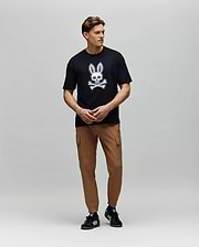 A man stands against a plain background wearing a black Psycho Bunny T-shirt with a white bunny and crossbones, MENS LLOYD CARGO JOGGER - B6P159B200 pants, and black athletic sneakers with white stripes.