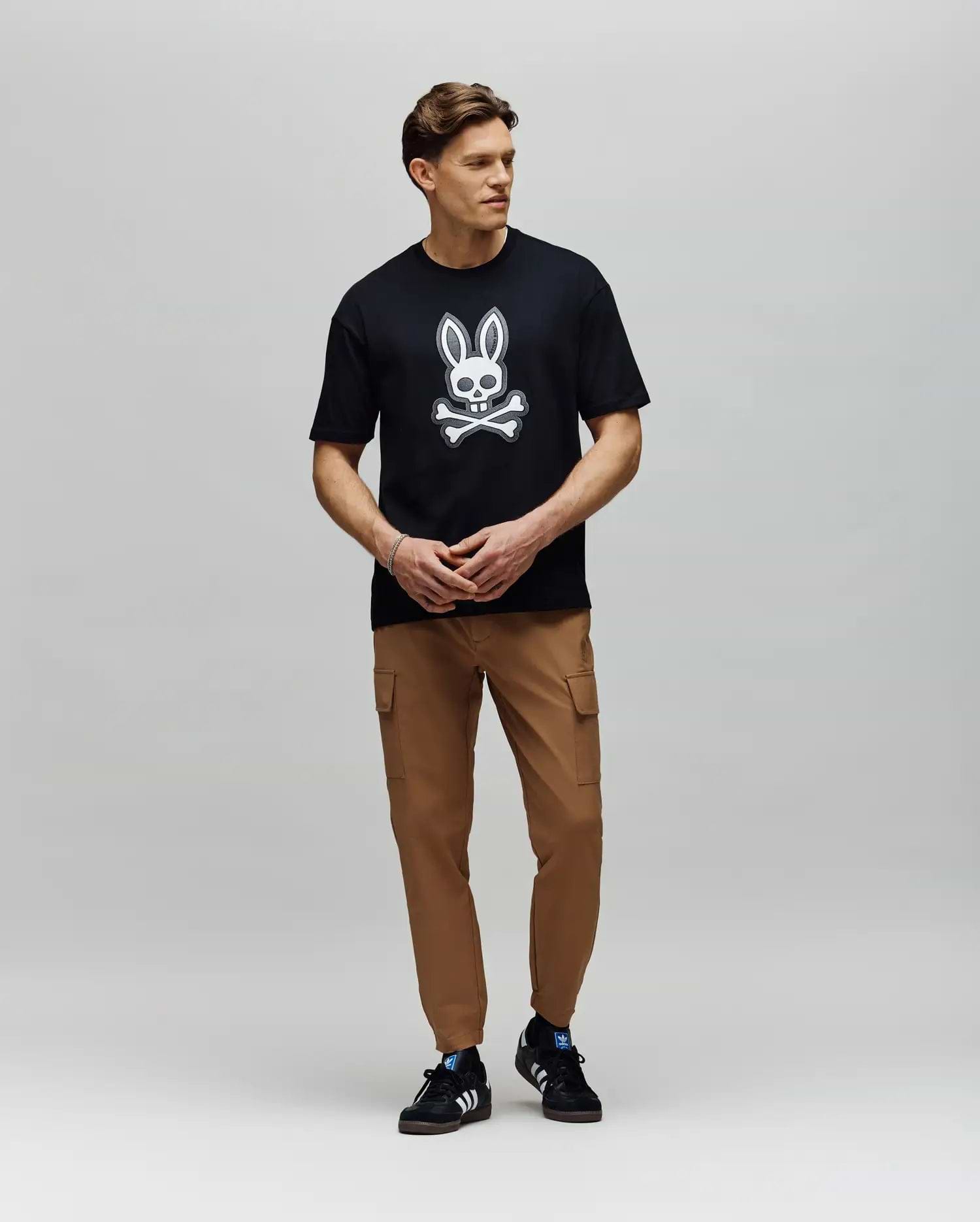 A man stands against a plain background wearing a black Psycho Bunny T-shirt with a white bunny and crossbones, MENS LLOYD CARGO JOGGER - B6P159B200 pants, and black athletic sneakers with white stripes.