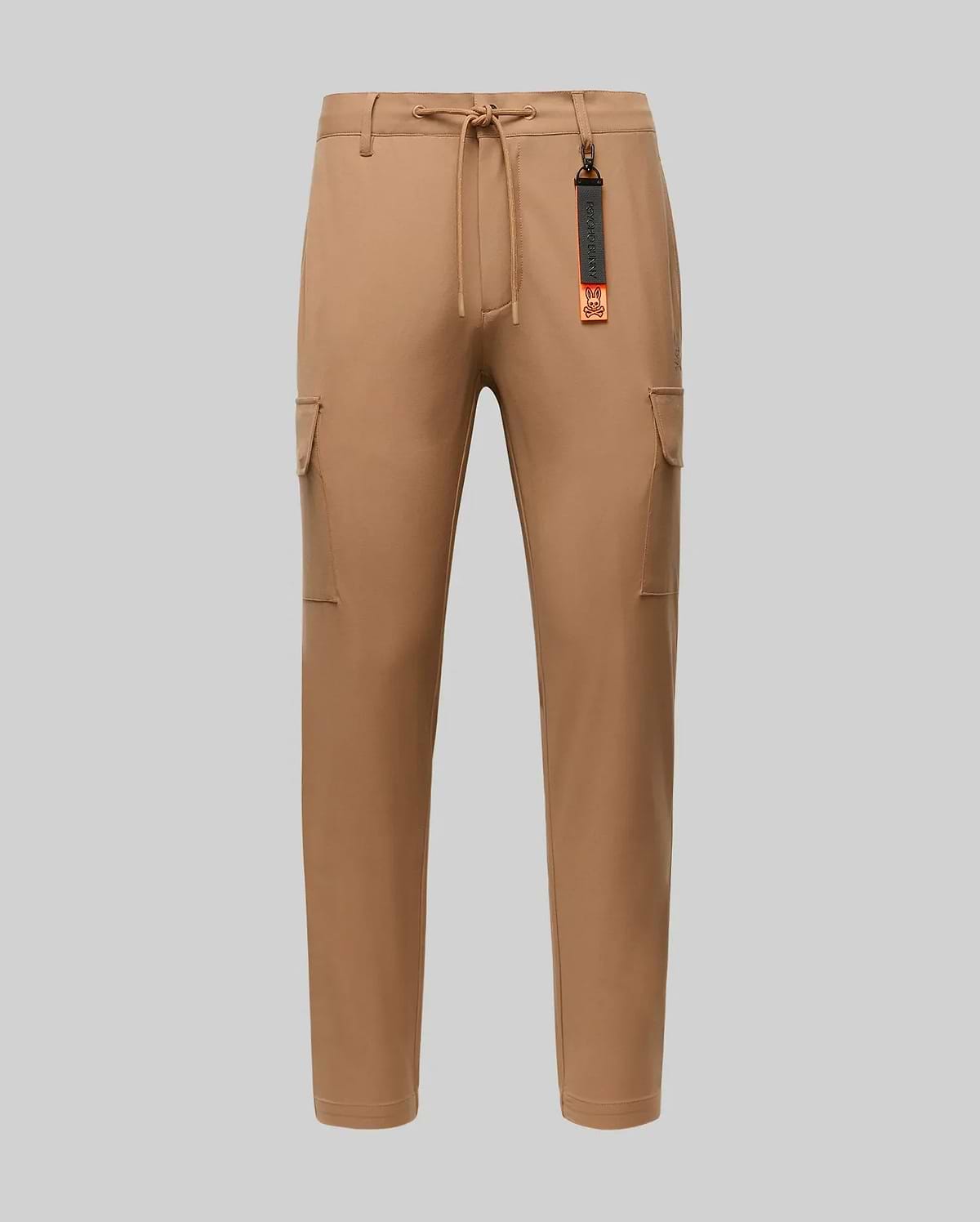 The Psycho Bunny MENS LLOYD CARGO JOGGER - B6P159B200 features tan fabric, elastic waistband, drawstring, side pockets, and a black-and-orange tag on a belt loop—perfect for upgrading your men's pants collection. Shown on a plain light gray background.
