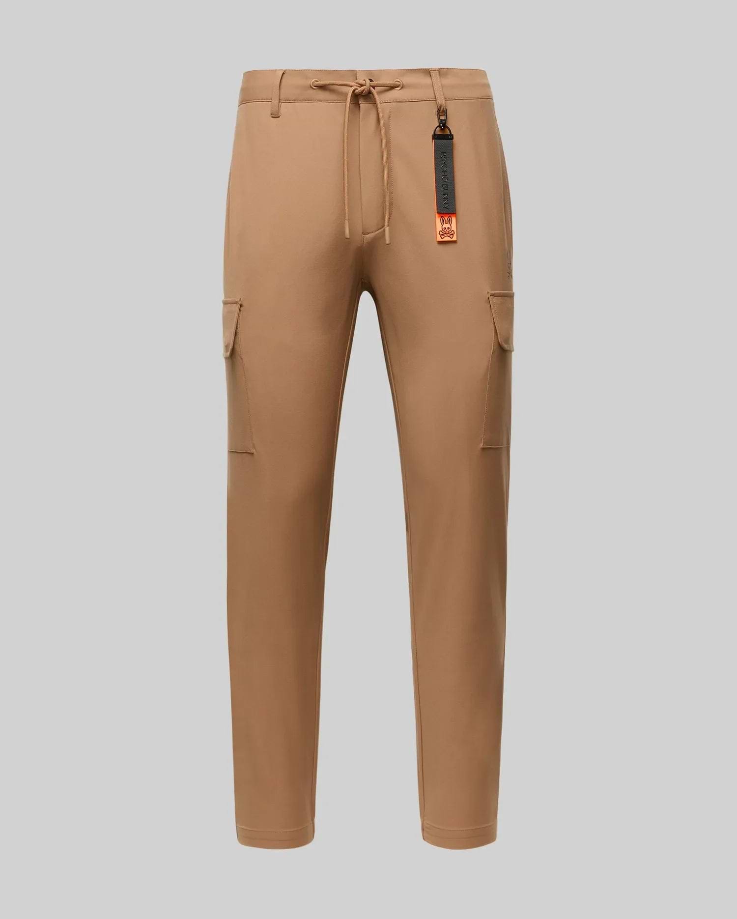 The Psycho Bunny MENS LLOYD CARGO JOGGER - B6P159B200 features tan fabric, elastic waistband, drawstring, side pockets, and a black-and-orange tag on a belt loop—perfect for upgrading your men's pants collection. Shown on a plain light gray background.