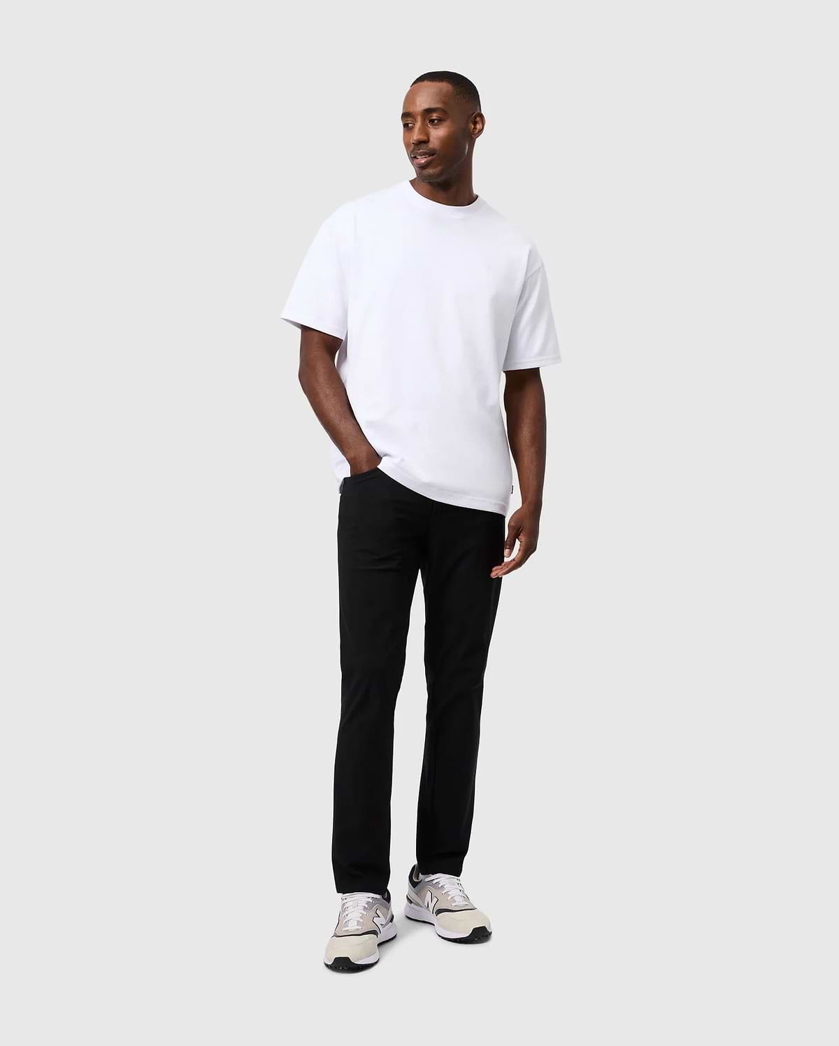 A person wearing a white t-shirt, the Psycho Bunny Mens Maverick 10K Pant - B6P788D200 with four-way stretch, and white sneakers stands against a plain gray background, looking slightly to the side with hands in pockets.
