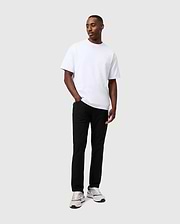 A person wearing a white t-shirt, the Psycho Bunny Mens Maverick 10K Pant - B6P788D200 with four-way stretch, and white sneakers stands against a plain gray background, looking slightly to the side with hands in pockets.