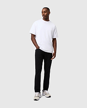 A person wearing a white t-shirt, the Psycho Bunny Mens Maverick 10K Pant - B6P788D200 with four-way stretch, and white sneakers stands against a plain gray background, looking slightly to the side with hands in pockets.