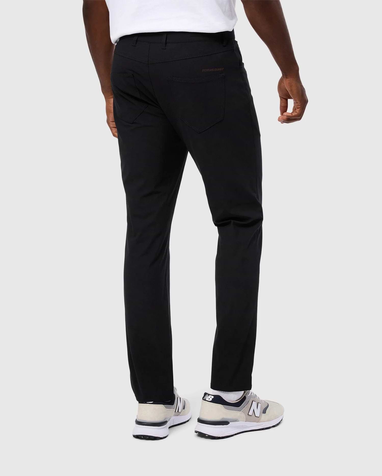 A person wearing Psycho Bunny's MENS MAVERICK 10K PANT - B6P788D200, showcasing a slim fit and five-pocket design, stands against a plain background. The black pants enhance the appearance of their white t-shirt, and their white and gray sneakers add a touch of subtle flair to complete the outfit.
