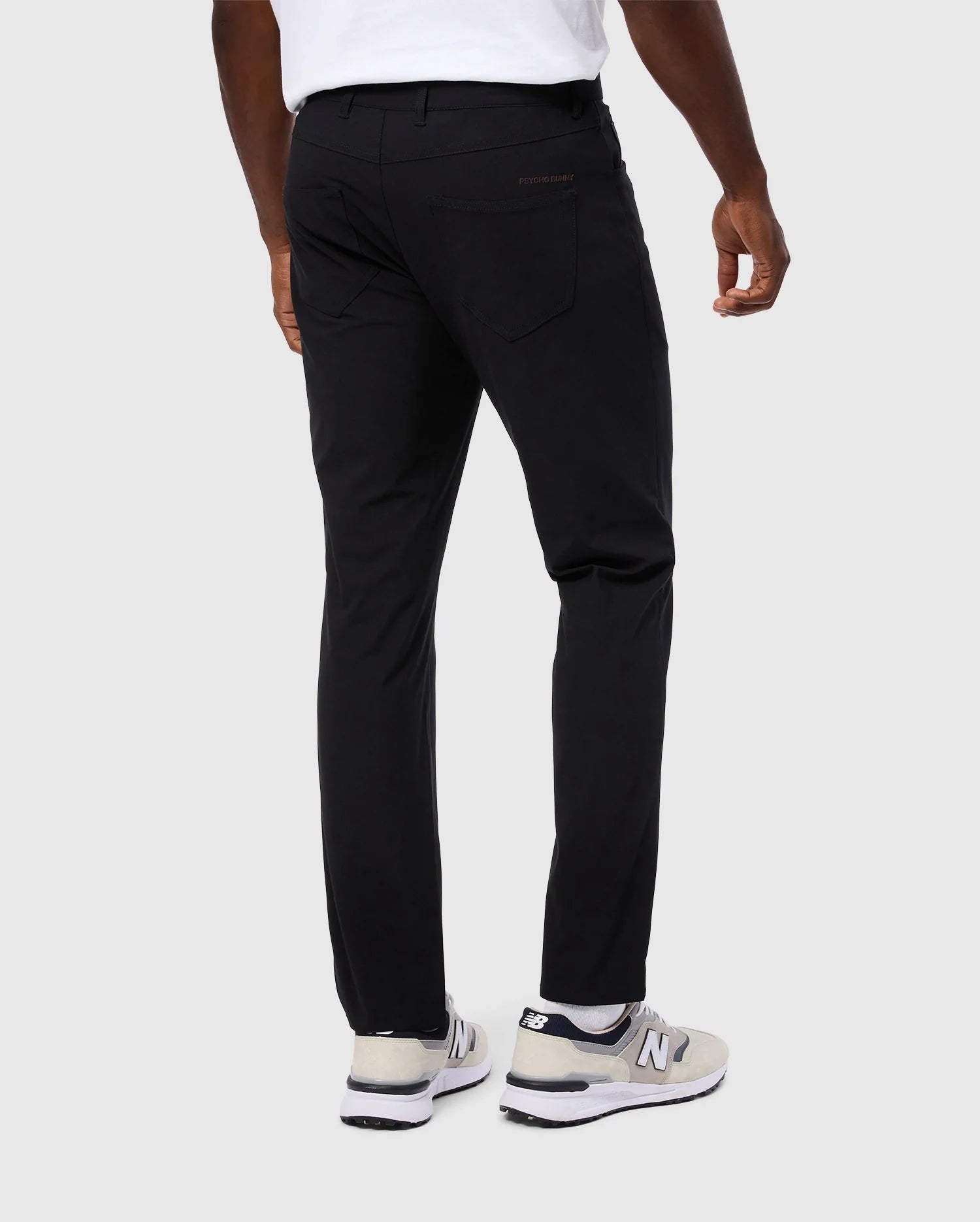 A person wearing Psycho Bunny's MENS MAVERICK 10K PANT - B6P788D200, showcasing a slim fit and five-pocket design, stands against a plain background. The black pants enhance the appearance of their white t-shirt, and their white and gray sneakers add a touch of subtle flair to complete the outfit.