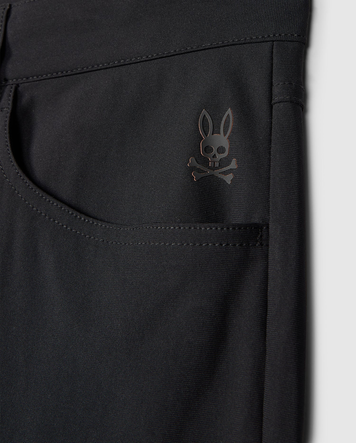 Close-up of the Psycho Bunny MENS MAVERICK 10K PANT - B6P788D200 in black highlighting a pocket adorned with a subtle skull and crossbones logo topped with bunny ears near the upper corner. The fabric looks smooth, slightly glossy, and incorporates four-way stretch for enhanced comfort.