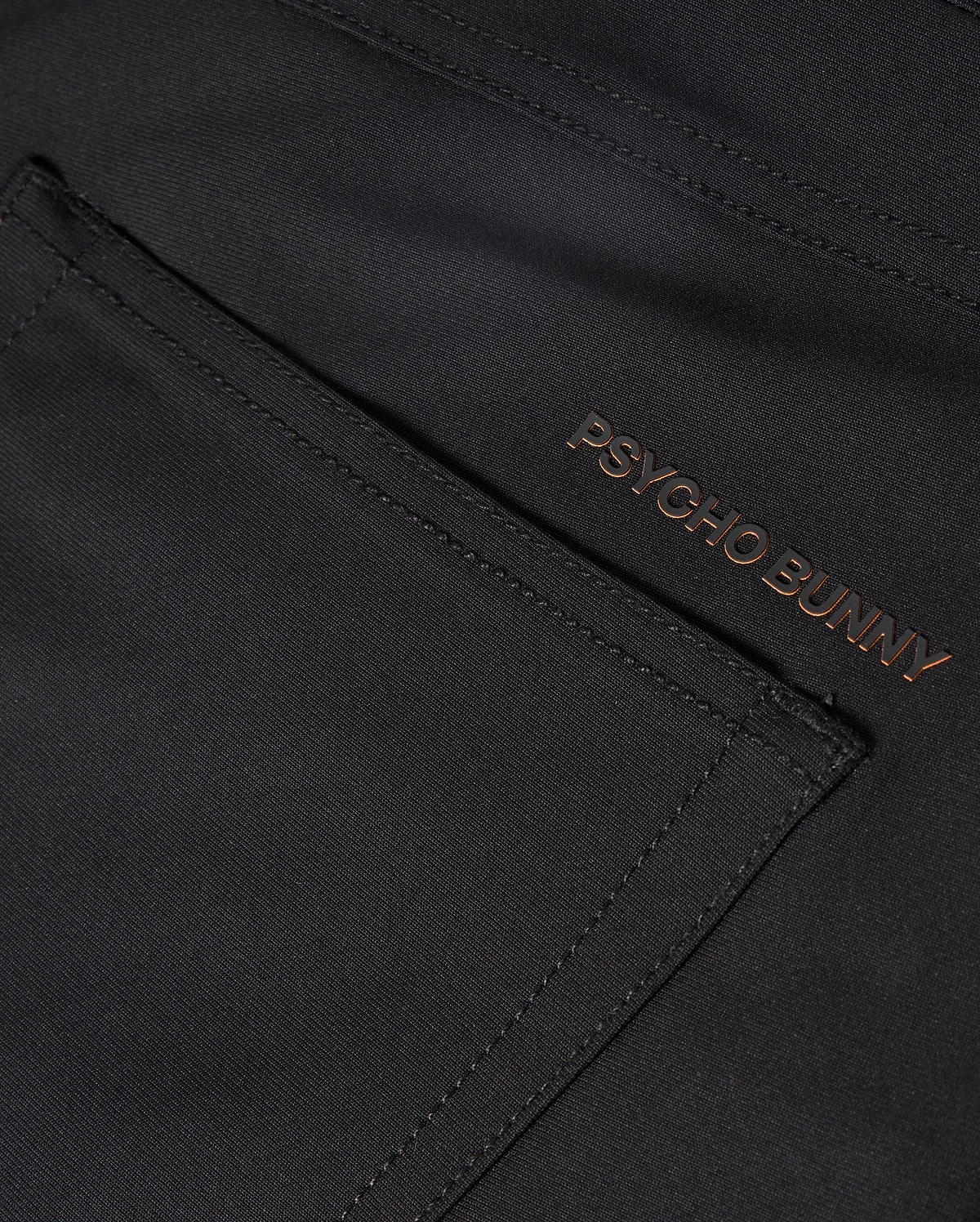 The MENS MAVERICK 10K PANT - B6P788D200 from Psycho Bunny showcases a sleek pocket in a close-up of the dark fabric. The brand name "PSYCHO BUNNY" is embroidered in a contrasting color near the edge, enhancing its modern design with four-way stretch for ultimate comfort.
