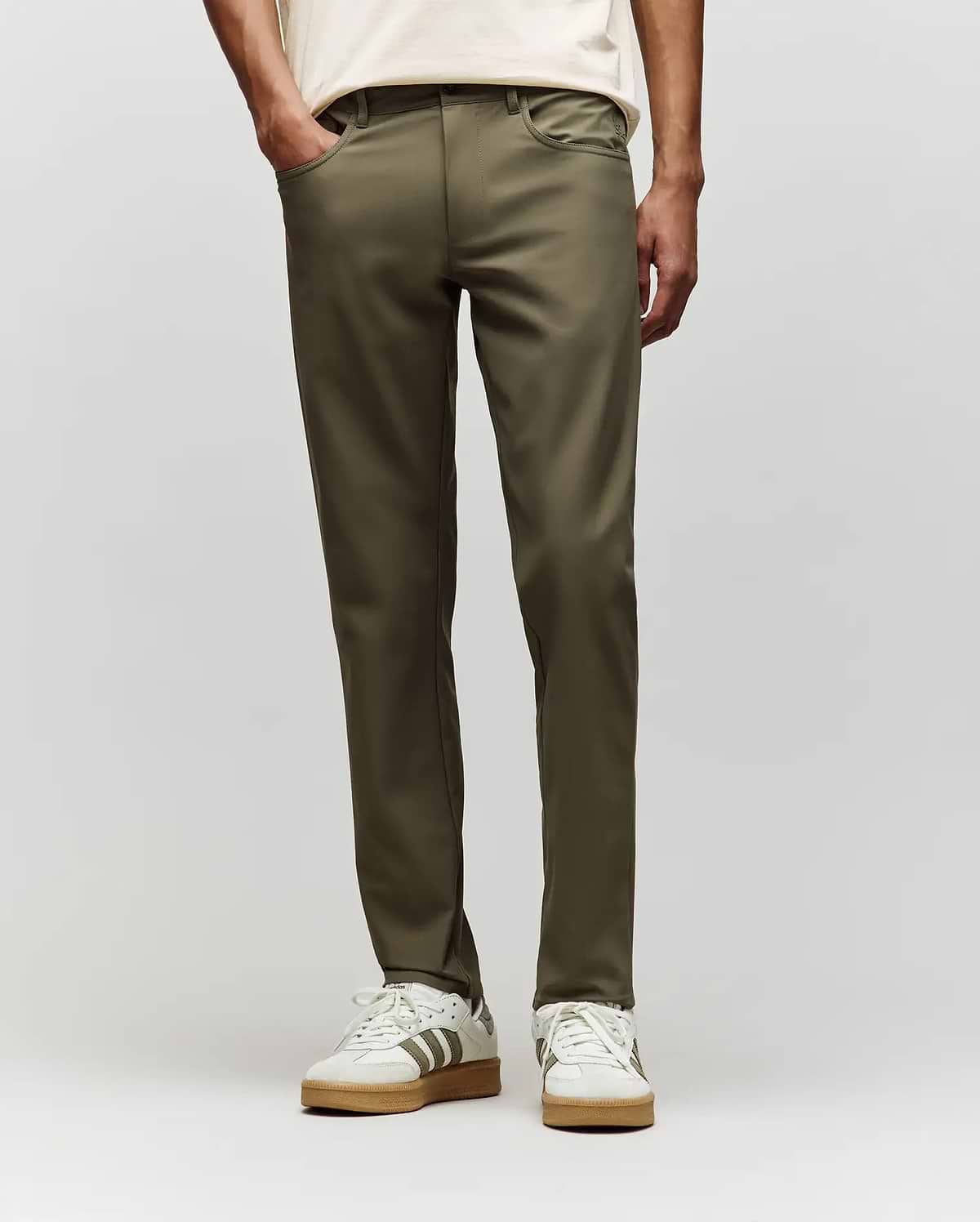 A person stands hands-in-pockets against a plain light background, wearing Psycho Bunny MENS MAVERICK 10K STEP PANTS - B6P788D200 in olive green, paired with a cream shirt and white sneakers with gray stripes. (1)