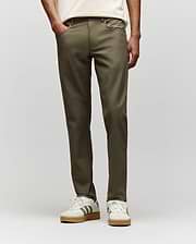 A person stands hands-in-pockets against a plain light background, wearing Psycho Bunny MENS MAVERICK 10K STEP PANTS - B6P788D200 in olive green, paired with a cream shirt and white sneakers with gray stripes. (1)