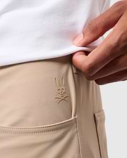 Close-up of a person wearing beige MENS MAVERICK 10K PANTS - B6P788D200 by Psycho Bunny, featuring a small, embroidered logo of a rabbit skull and crossbones above the front pocket. The person is also wearing a white shirt tucked in and is slightly lifting it to reveal the logo.