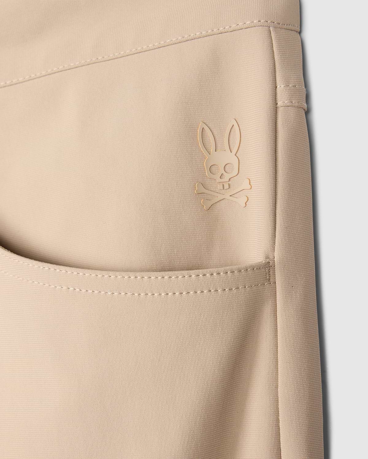 Close-up of a beige Psycho Bunny MENS MAVERICK 10K PANT - B6P788D200 pocket featuring an embroidered logo of a rabbit with crossbones underneath. The stitching and texture of the fabric, along with its four-way stretch, are clearly visible, emphasizing the detailed design and craftsmanship.