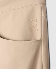 Close-up of a beige Psycho Bunny MENS MAVERICK 10K PANT - B6P788D200 pocket featuring an embroidered logo of a rabbit with crossbones underneath. The stitching and texture of the fabric, along with its four-way stretch, are clearly visible, emphasizing the detailed design and craftsmanship.