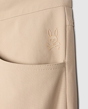 Close-up of a beige Psycho Bunny MENS MAVERICK 10K PANT - B6P788D200 pocket featuring an embroidered logo of a rabbit with crossbones underneath. The stitching and texture of the fabric, along with its four-way stretch, are clearly visible, emphasizing the detailed design and craftsmanship.