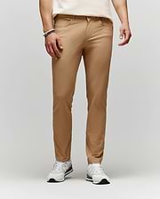 A person wears Psycho Bunny MENS MAVERICK 10K STEP PANT - B6P788D200 in tan, styled with a cream t-shirt, white sneakers, and bracelets, standing against a plain light gray background with their upper body partially out of frame.