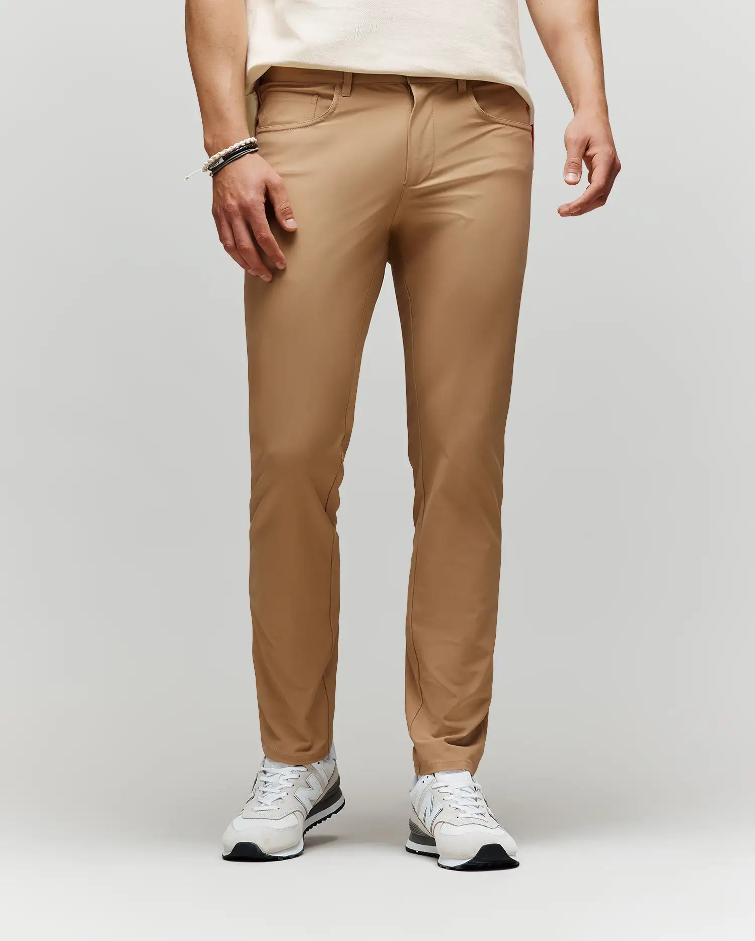 A person wears Psycho Bunny MENS MAVERICK 10K STEP PANT - B6P788D200 in tan, styled with a cream t-shirt, white sneakers, and bracelets, standing against a plain light gray background with their upper body partially out of frame.