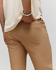 A person wearing Psycho Bunny’s light brown MENS MAVERICK 10K STEP PANT (B6P788D200) with five-pocket styling, decorative seam, and "PEYOK BURY" on the pocket stands with one hand in their back pocket, paired with a cream shirt and white bracelet.