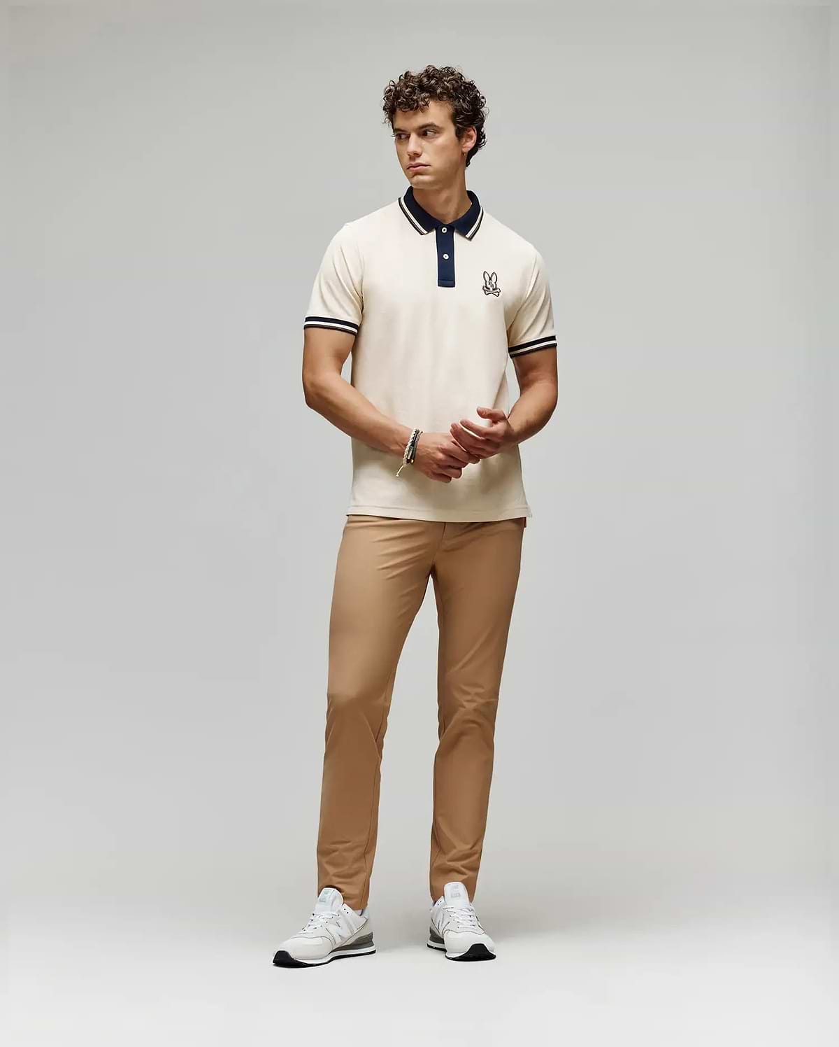 A man stands against a plain background wearing a cream polo with dark collar and sleeve trim, Psycho Bunny MENS MAVERICK 10K STEP PANT - B6P788D200 with five-pocket styling, and white sneakers. He clasps his hands in front and looks to the side.