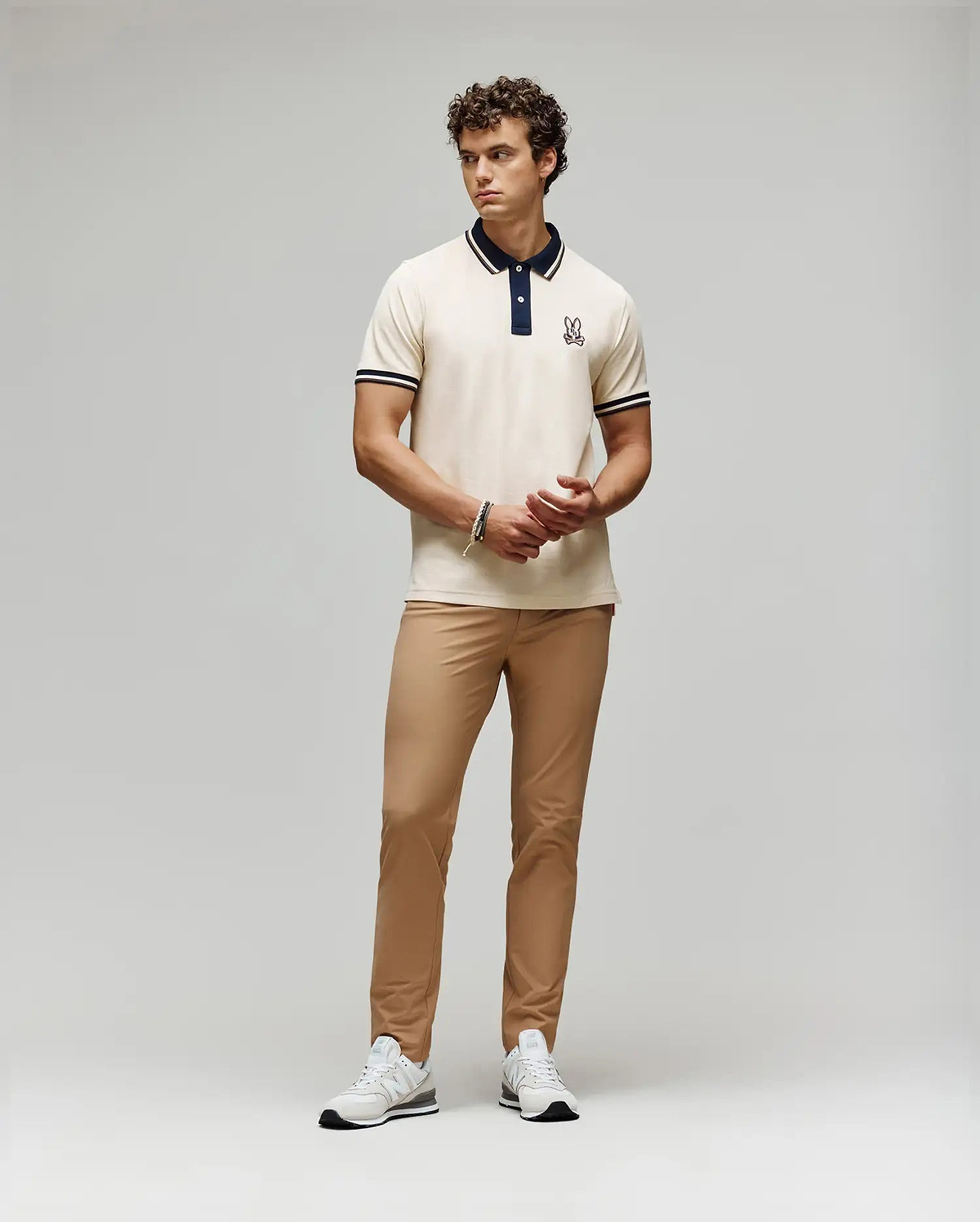 A man stands against a plain background wearing a cream polo with dark collar and sleeve trim, Psycho Bunny MENS MAVERICK 10K STEP PANT - B6P788D200 with five-pocket styling, and white sneakers. He clasps his hands in front and looks to the side.
