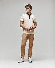 A man stands against a plain background wearing a cream polo with dark collar and sleeve trim, Psycho Bunny MENS MAVERICK 10K STEP PANT - B6P788D200 with five-pocket styling, and white sneakers. He clasps his hands in front and looks to the side.