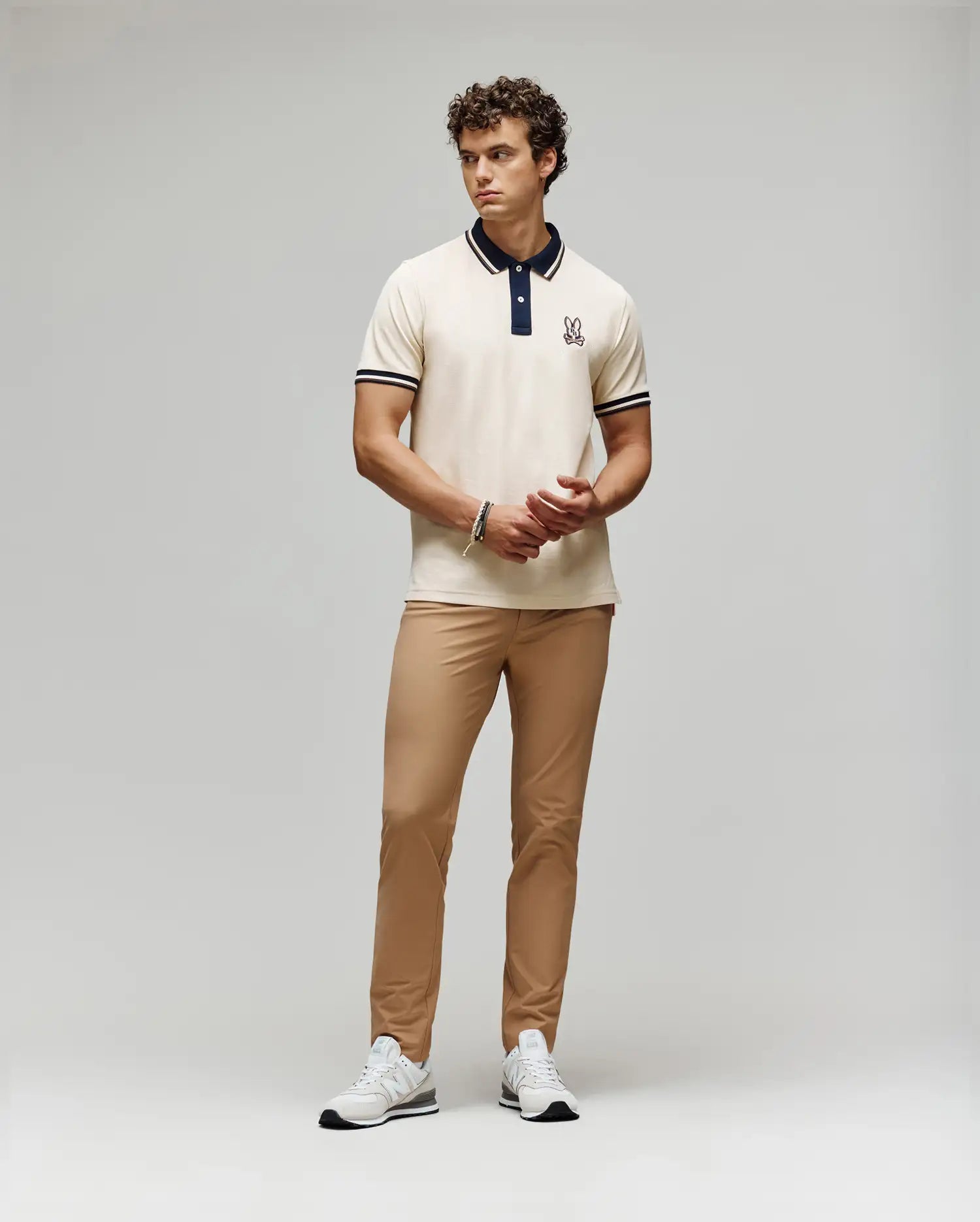 A man stands against a plain background wearing a cream polo with dark collar and sleeve trim, Psycho Bunny MENS MAVERICK 10K STEP PANT - B6P788D200 with five-pocket styling, and white sneakers. He clasps his hands in front and looks to the side.