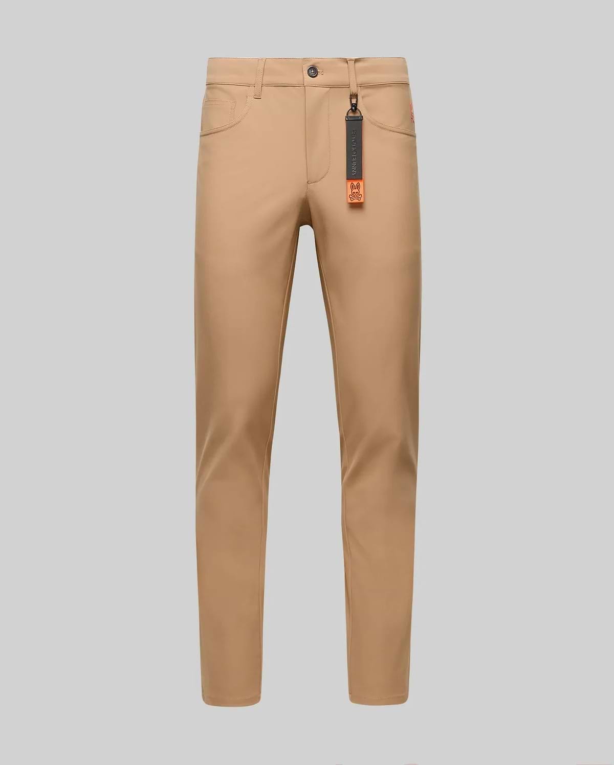 The Psycho Bunny MENS MAVERICK 10K STEP PANT - B6P788D200 is shown in slim-fit tan, featuring four-way stretch, front and back pockets, belt loops, and a black tag with an orange accent on the front belt loop against a gray background.