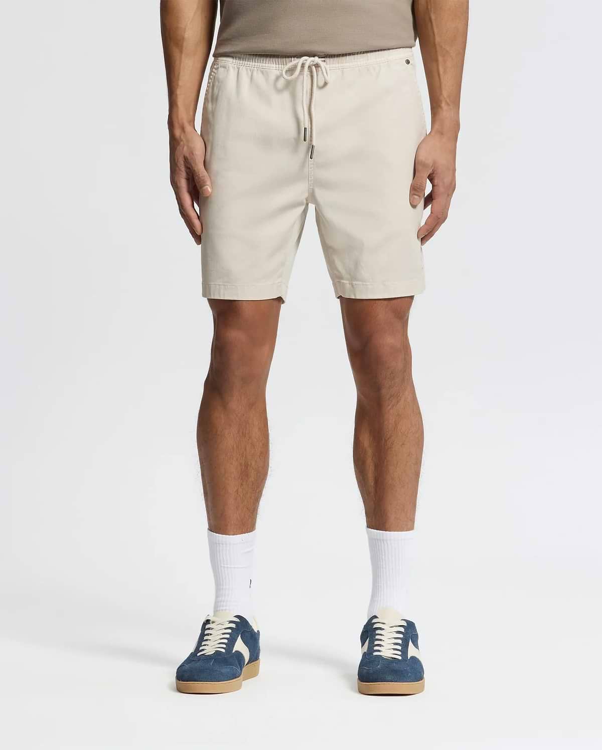 Effortlessly stylish, a person wears beige Psycho Bunny MENS WILLIS STRETCH TENCEL SHORTS (B6R584F200) with an elastic waistband, a taupe tank top, white socks, and blue sneakers against a plain white background.