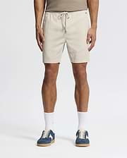 Effortlessly stylish, a person wears beige Psycho Bunny MENS WILLIS STRETCH TENCEL SHORTS (B6R584F200) with an elastic waistband, a taupe tank top, white socks, and blue sneakers against a plain white background.
