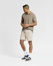 Wearing a tan polo shirt, Psycho Bunny's Men's Willis Stretch Tencel Shorts in light beige with an elasticized waistband, white socks, and blue sneakers, a man stands stylishly against a plain white background, gazing right with his hands at his sides.