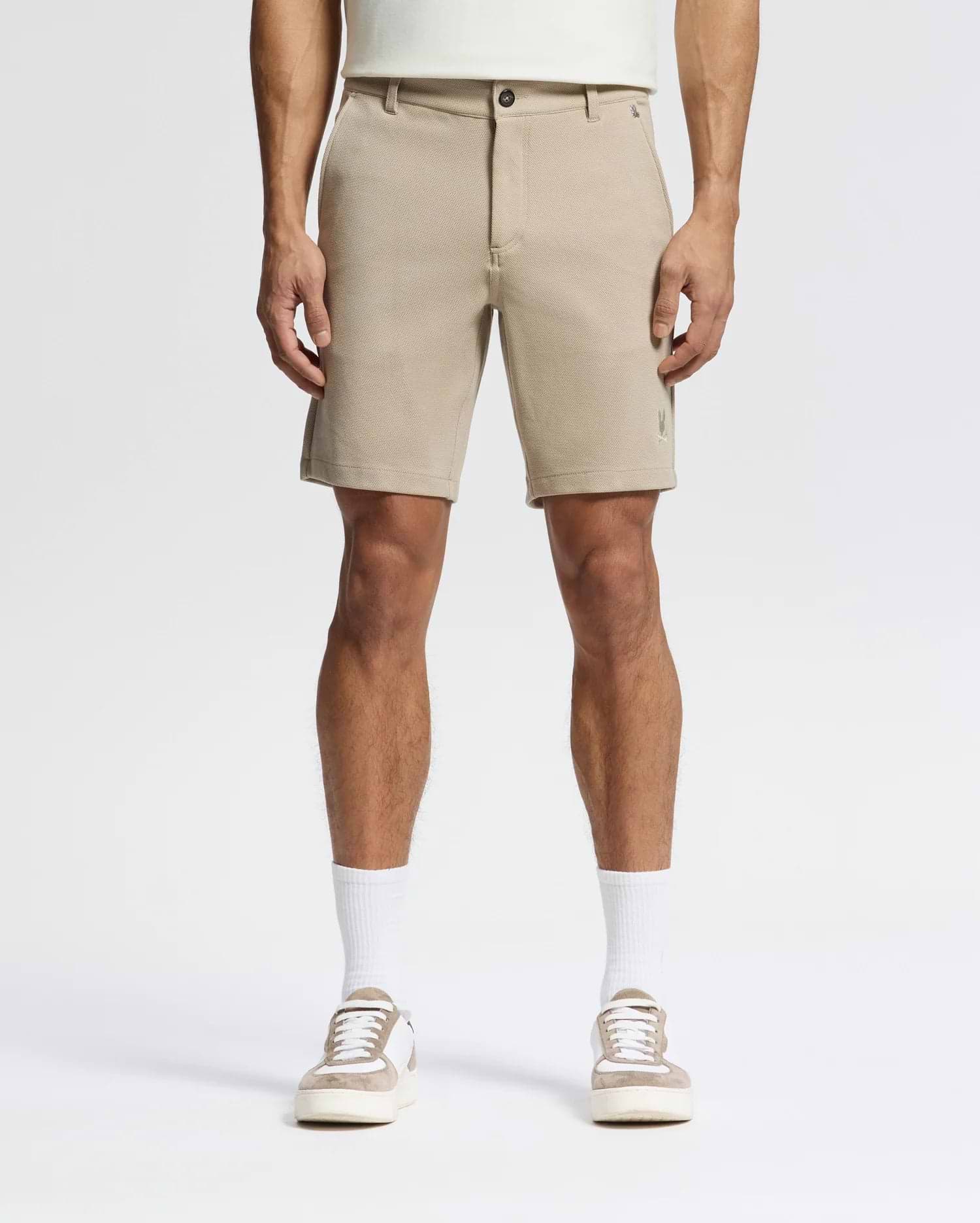 A person wears Psycho Bunny MENS SHIRO KNIT HONEYCOMB SHORTS (B6R585F200), white crew socks, and beige-and-white sneakers, facing forward against a plain white background. The photo is cropped at the torso to show only the lower half.