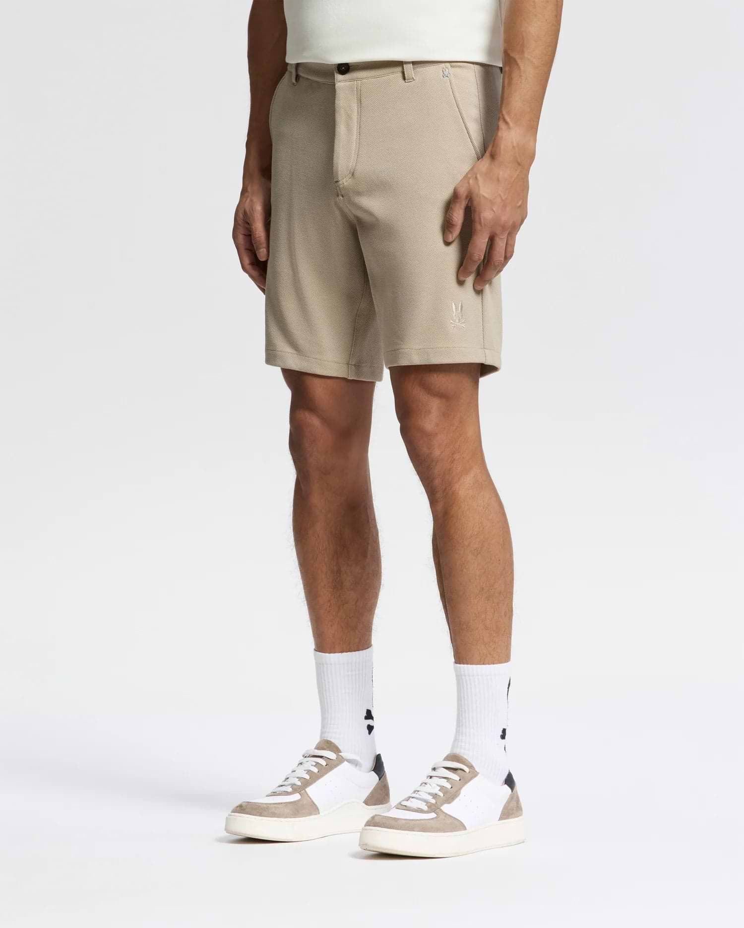 A man models Psycho Bunny’s MENS SHIRO KNIT HONEYCOMB SHORT - B6R585F200 in beige, paired with logo white socks and white sneakers with beige accents, standing against a white background for a stylish, versatile look.