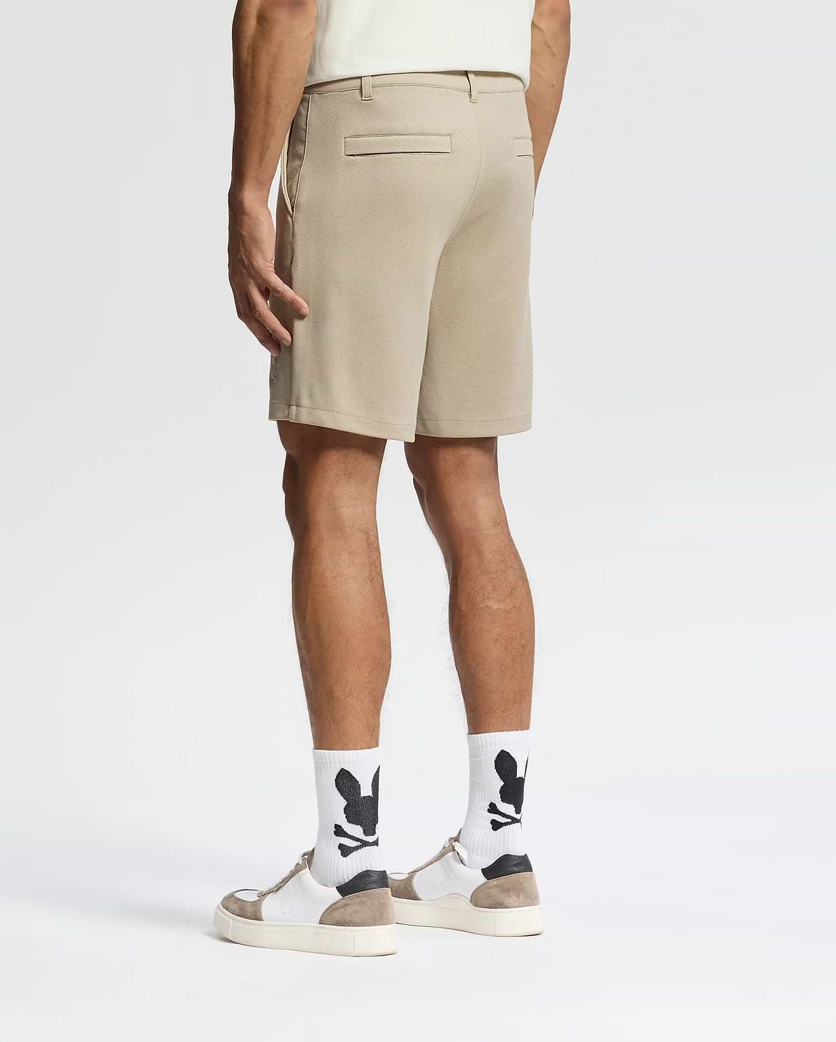 A man, back turned, wears Psycho Bunny MENS SHIRO KNIT HONEYCOMB SHORTS (B6R585F200), white socks with black graphics, white sneakers with tan accents, and a light shirt against a plain white background for a smart-casual look.