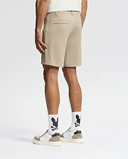 A man, back turned, wears Psycho Bunny MENS SHIRO KNIT HONEYCOMB SHORTS (B6R585F200), white socks with black graphics, white sneakers with tan accents, and a light shirt against a plain white background for a smart-casual look.