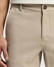 A close-up of a person wearing Psycho Bunny MENS SHIRO KNIT HONEYCOMB SHORTS (B6R585F200) in beige with a honeycomb piqué pattern, black button, white shirt tucked in, and their left hand at their side. Subtle woven texture shown.