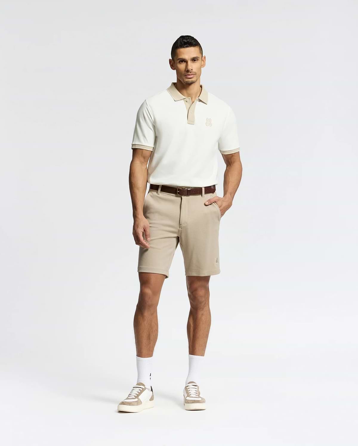 A man stands against a white background wearing Psycho Bunny's MENS SHIRO KNIT HONEYCOMB SHORT (B6R585F200) with a brown belt, white crew socks, and beige sneakers. One hand is in his pocket as he faces forward with a neutral expression.