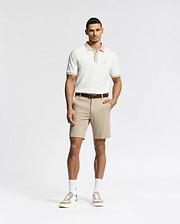A man stands against a white background wearing Psycho Bunny's MENS SHIRO KNIT HONEYCOMB SHORT (B6R585F200) with a brown belt, white crew socks, and beige sneakers. One hand is in his pocket as he faces forward with a neutral expression.
