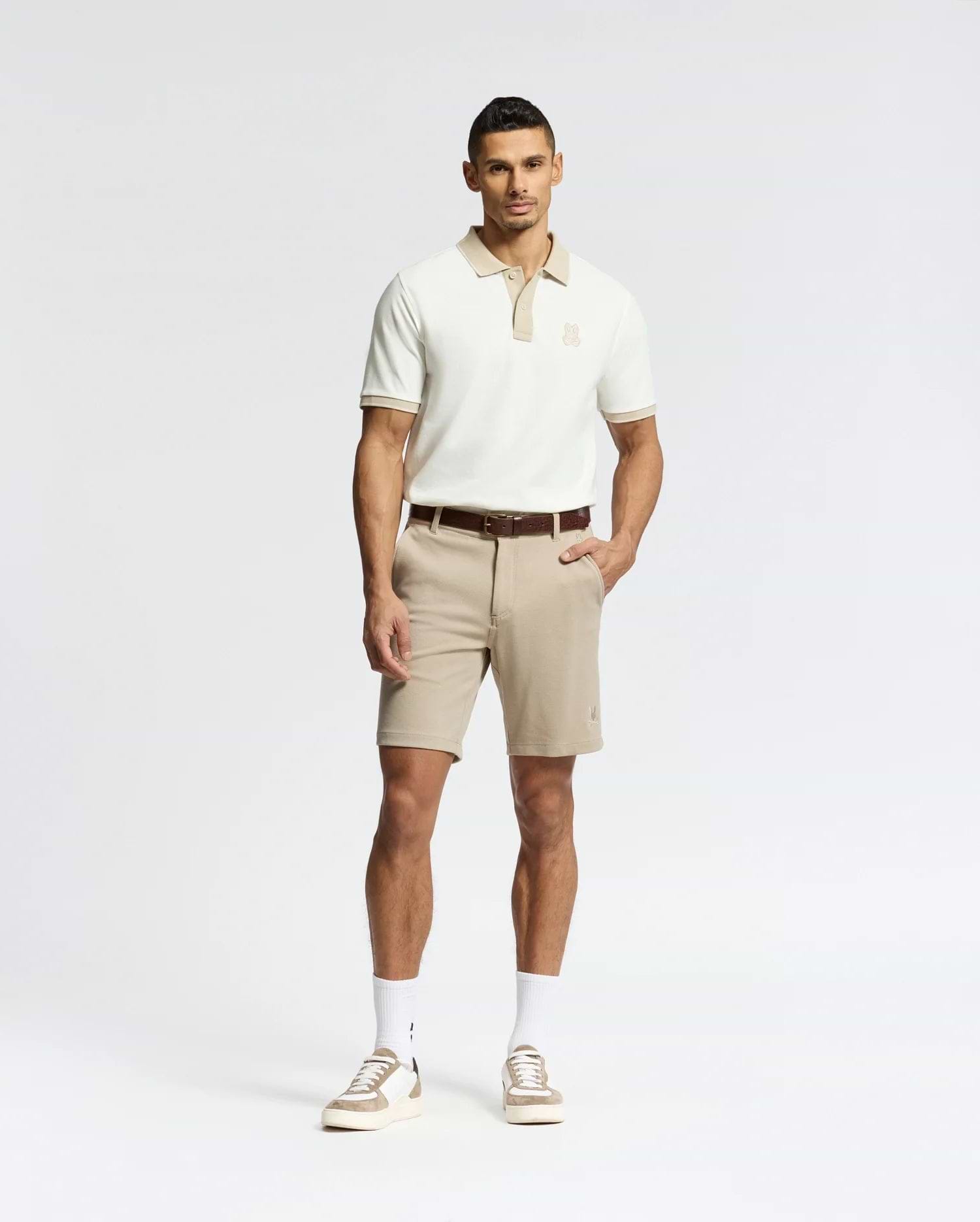 A man stands against a white background wearing Psycho Bunny's MENS SHIRO KNIT HONEYCOMB SHORT (B6R585F200) with a brown belt, white crew socks, and beige sneakers. One hand is in his pocket as he faces forward with a neutral expression.