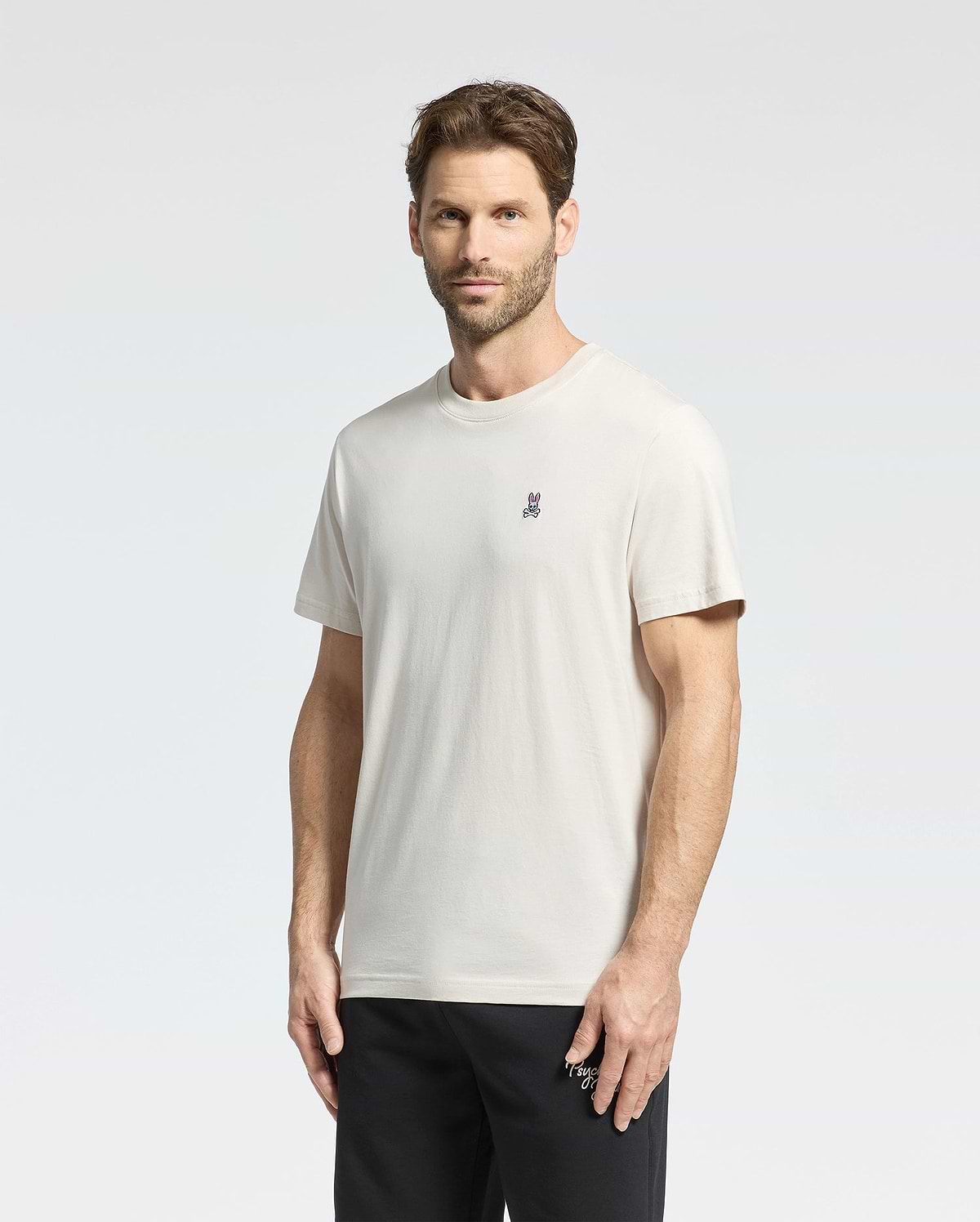 A man with short brown hair wears a Psycho Bunny Men's Classic Crew Neck Tee in premium jersey with a small embroidered chest logo, paired with black pants. He stands against a plain white background, looking at the camera neutrally.