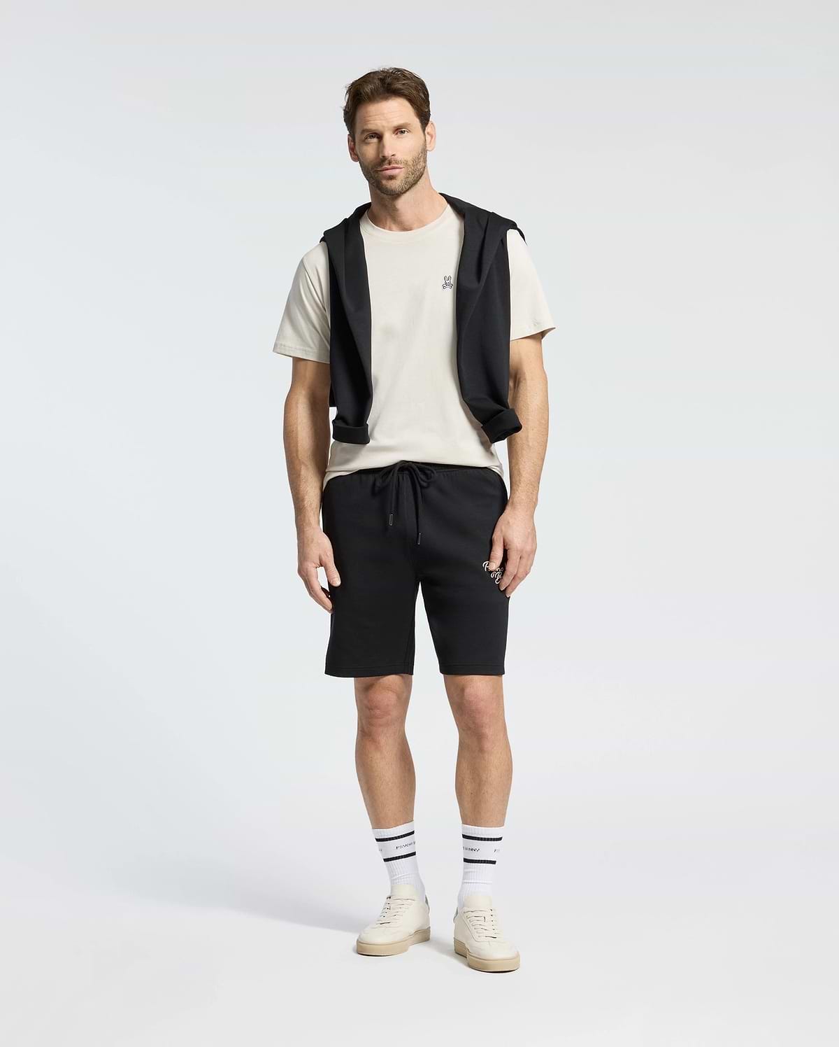 A man stands against a plain background wearing a Psycho Bunny MENS CLASSIC CREW NECK TEE (B6U014B200) in white, paired with black shorts and a black sweatshirt draped over his shoulders. His look is completed with striped socks and white sneakers.