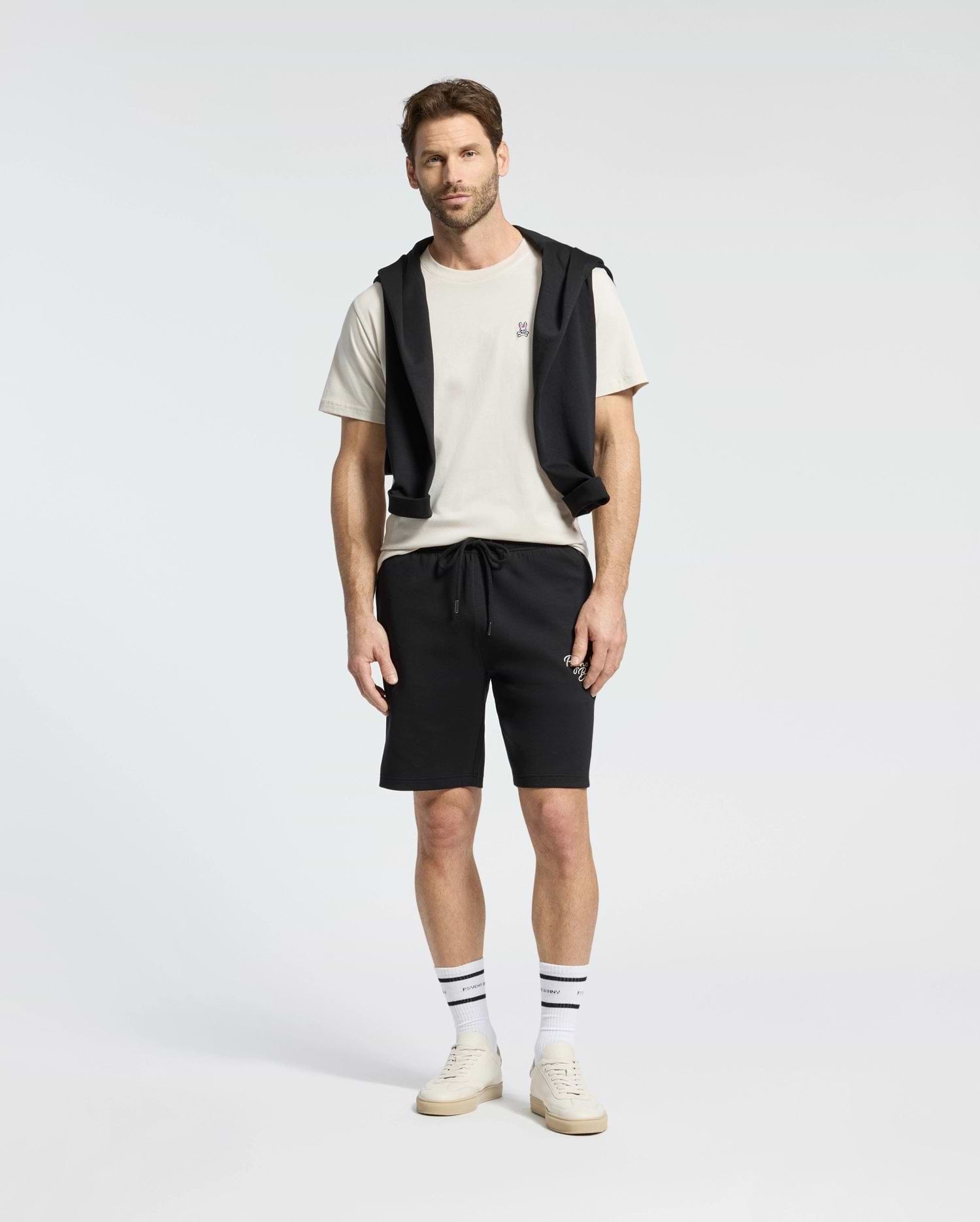 A man stands against a plain background wearing a Psycho Bunny MENS CLASSIC CREW NECK TEE (B6U014B200) in white, paired with black shorts and a black sweatshirt draped over his shoulders. His look is completed with striped socks and white sneakers.