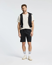 A man stands against a plain background wearing a Psycho Bunny MENS CLASSIC CREW NECK TEE (B6U014B200) in white, paired with black shorts and a black sweatshirt draped over his shoulders. His look is completed with striped socks and white sneakers.