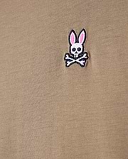 Close-up of a khaki tee made from 100% Pima cotton, showcasing the Psycho Bunny embroidered logo with a cartoonish skull and crossbones design featuring rabbit ears.