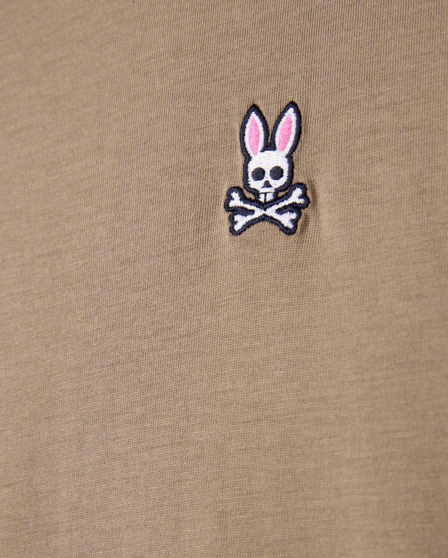 Close-up of a khaki tee made from 100% Pima cotton, showcasing the Psycho Bunny embroidered logo with a cartoonish skull and crossbones design featuring rabbit ears.