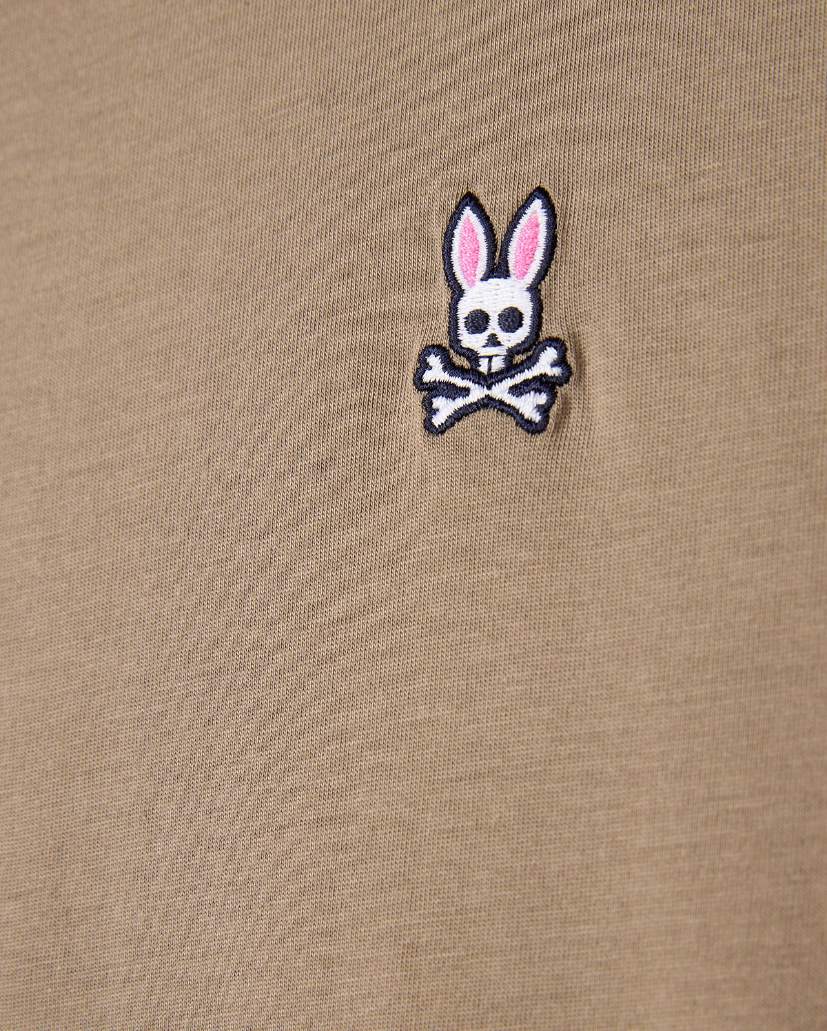 Close-up of a khaki tee made from 100% Pima cotton, showcasing the Psycho Bunny embroidered logo with a cartoonish skull and crossbones design featuring rabbit ears.