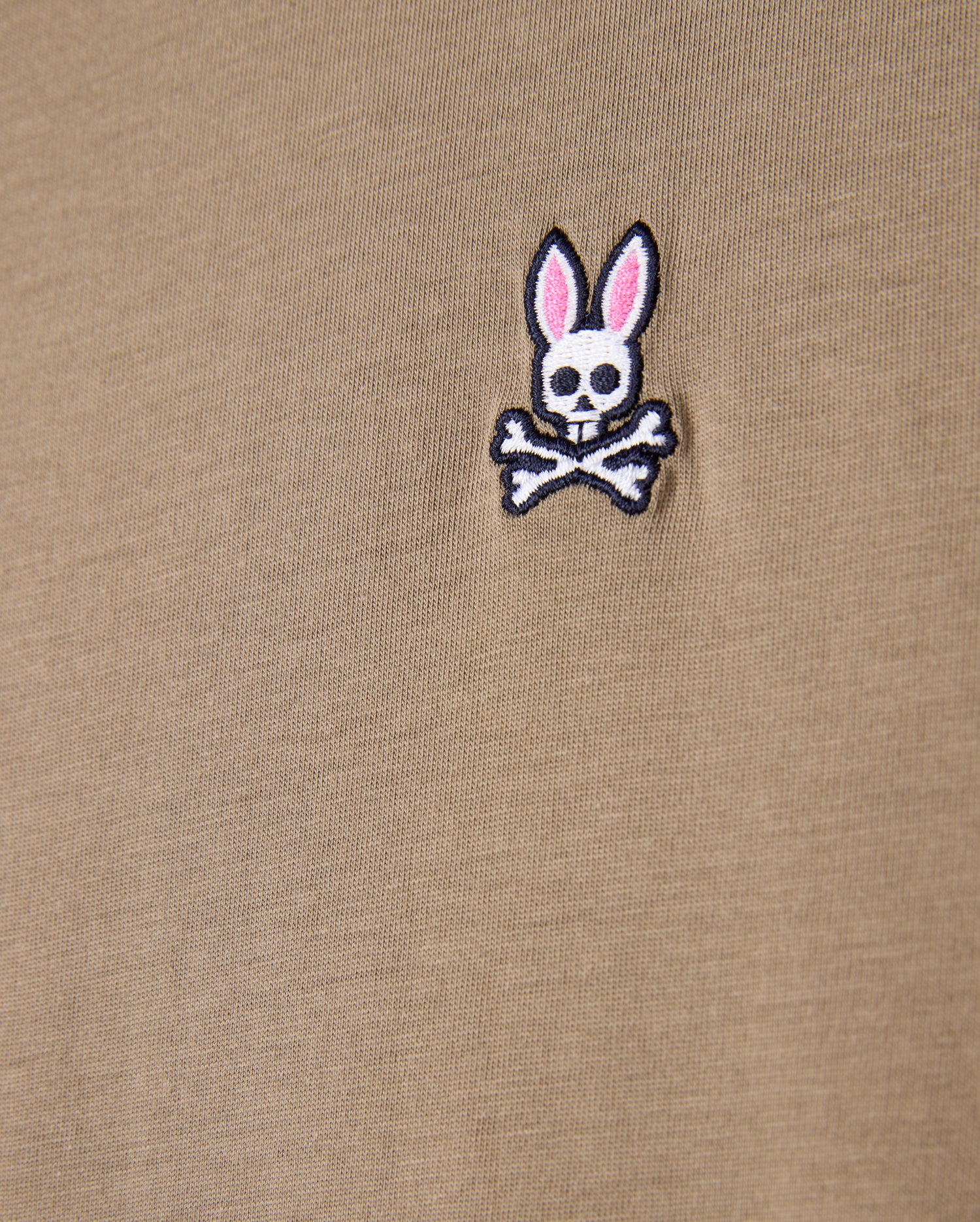 Close-up of a khaki tee made from 100% Pima cotton, showcasing the Psycho Bunny embroidered logo with a cartoonish skull and crossbones design featuring rabbit ears.