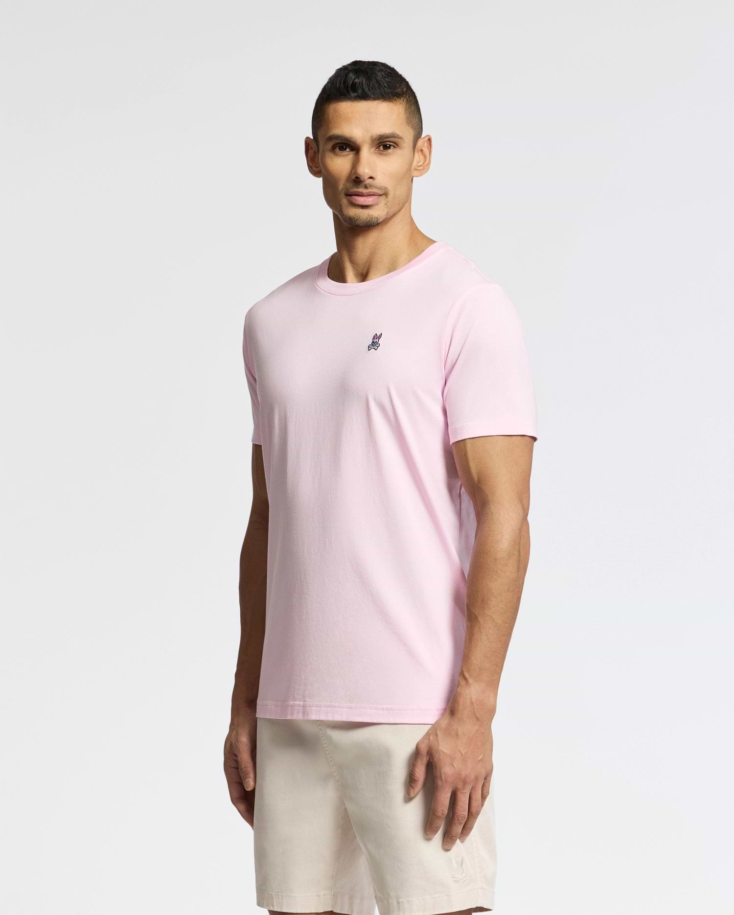 Against a plain backdrop, a person wears the Psycho Bunny Men's Classic Crew Neck Tee (B6U014CRPC) in light pink, made from soft pima cotton featuring a small chest emblem, paired with beige shorts. Its regular fit complements their slight smile as they gaze ahead.